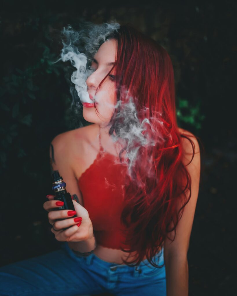 pexels-photo-13871866-13871866 Young woman with vibrant red hair uses a vape device, surrounded by smoke in an outdoor setting.